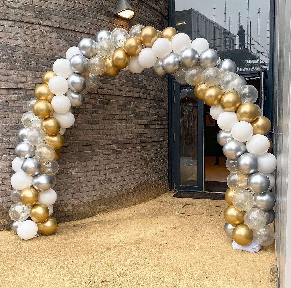 Un arco de globos dorados, plateados y blancos se encuentra frente a un edificio de ladrillos.