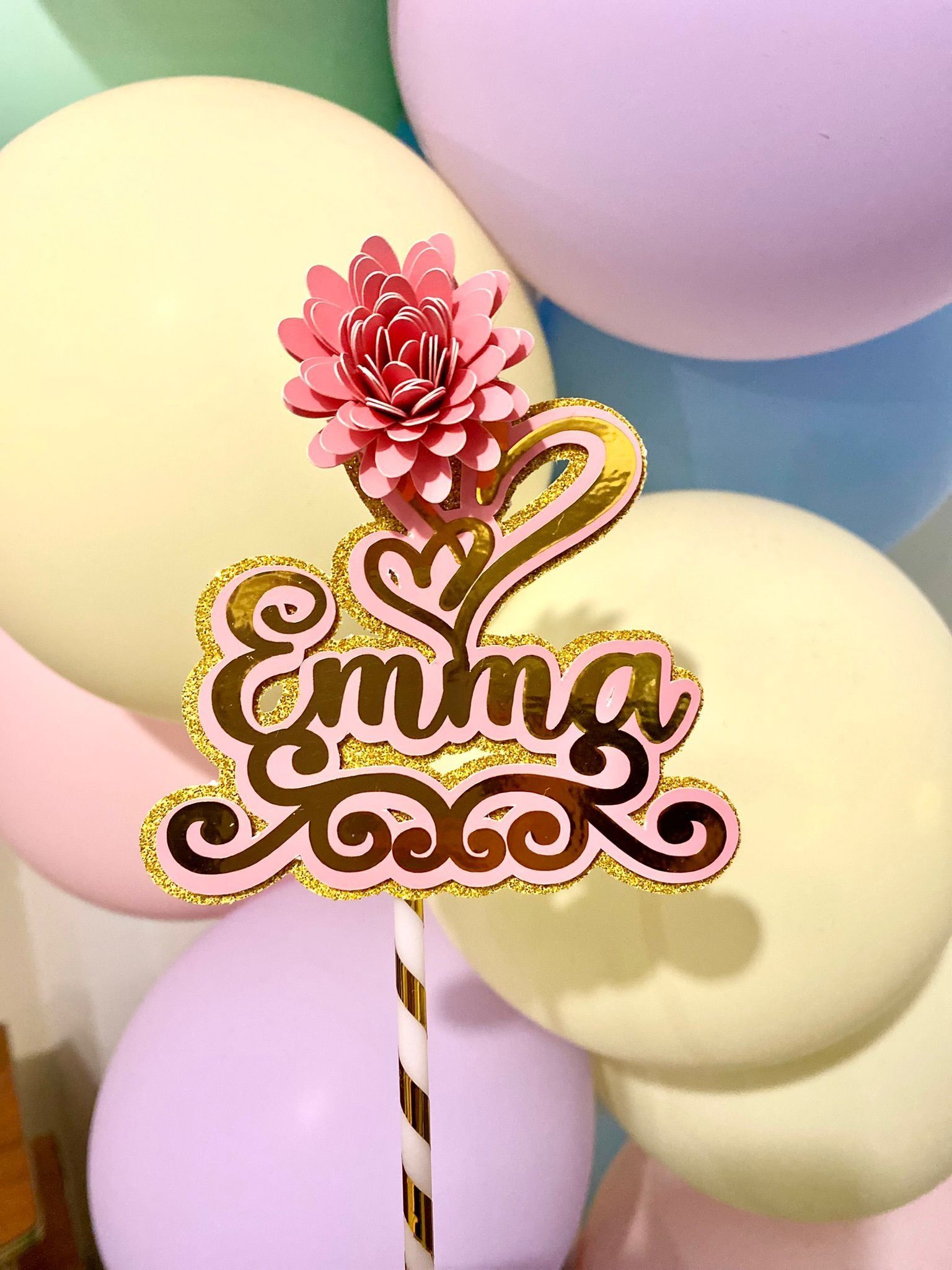 Un adorno para tarta con el nombre Emma está rodeado de globos.