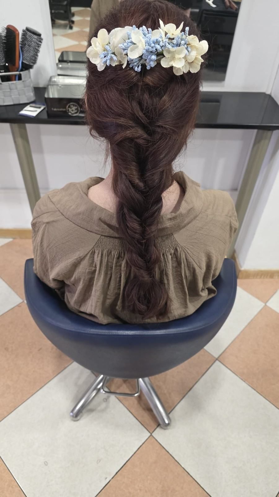 Mujer sentada en una silla de peluquería con el pelo trenzado y un tocado floral blanco y azul.