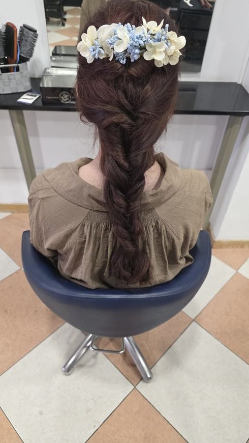Mujer sentada en una silla de peluquería con el pelo trenzado y un tocado floral blanco y azul.
