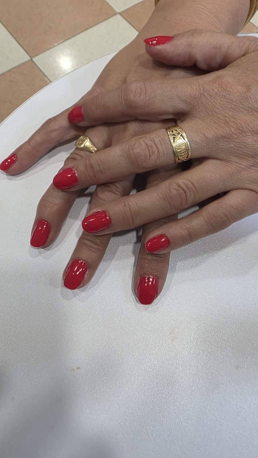 Dos manos con uñas de color rojo brillante y anillos de oro descansando sobre una encimera blanca.