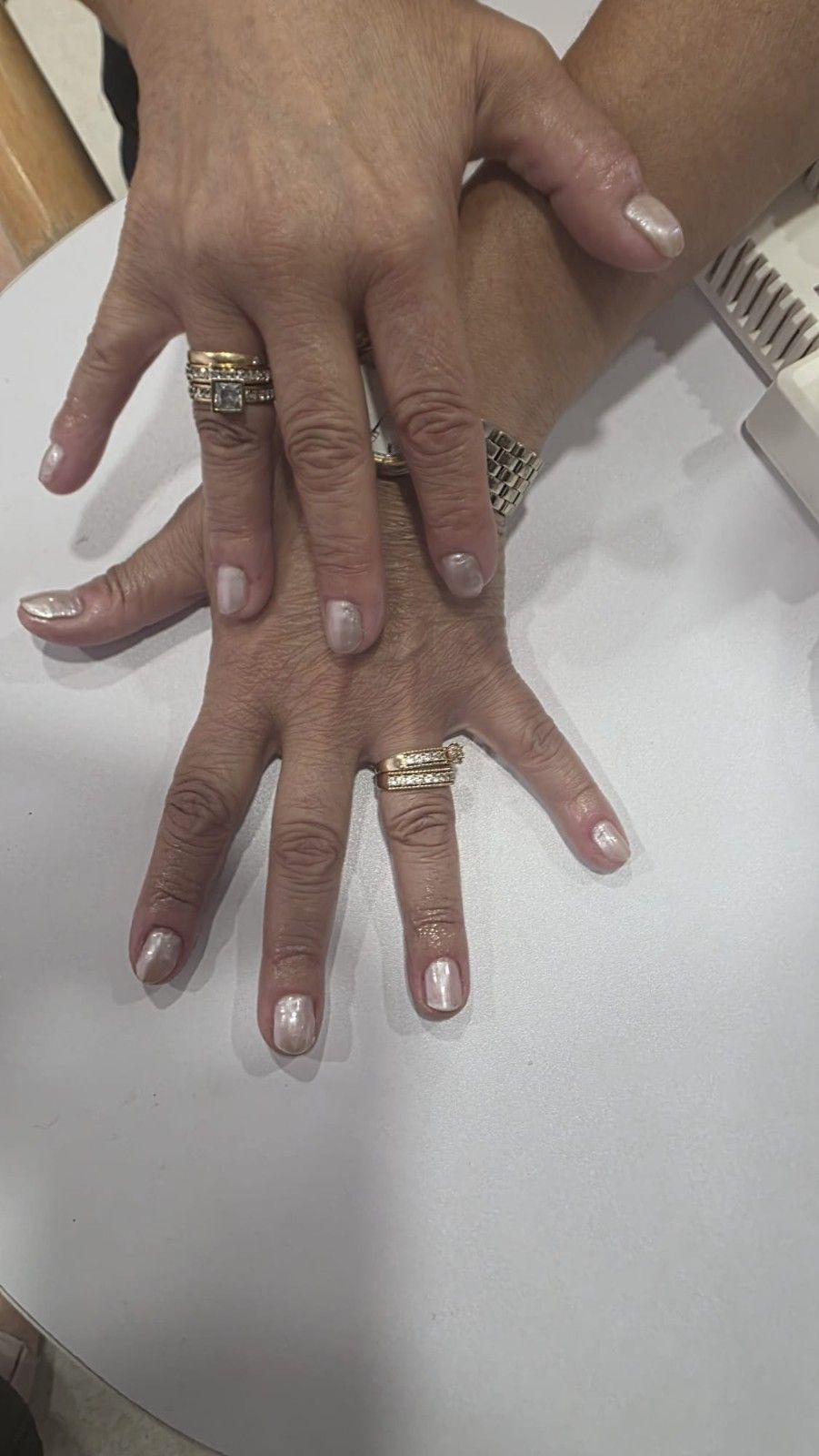 Dos manos apiladas sobre una mesa blanca, luciendo anillos de oro y esmalte de uñas rosa pálido.