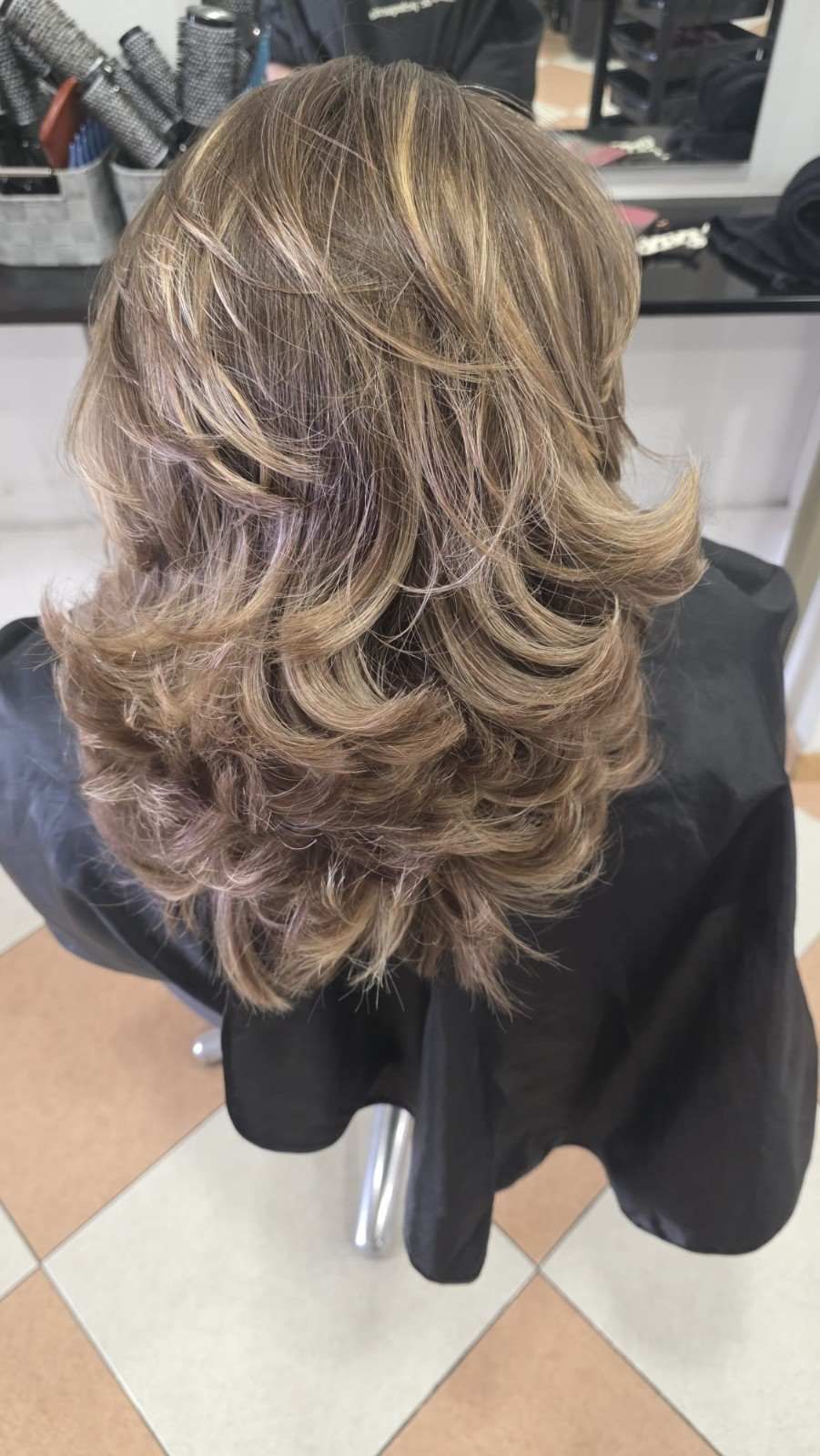 Cabello castaño a capas hasta los hombros con reflejos rubios, peinado en suaves rizos en una silla de peluquería.