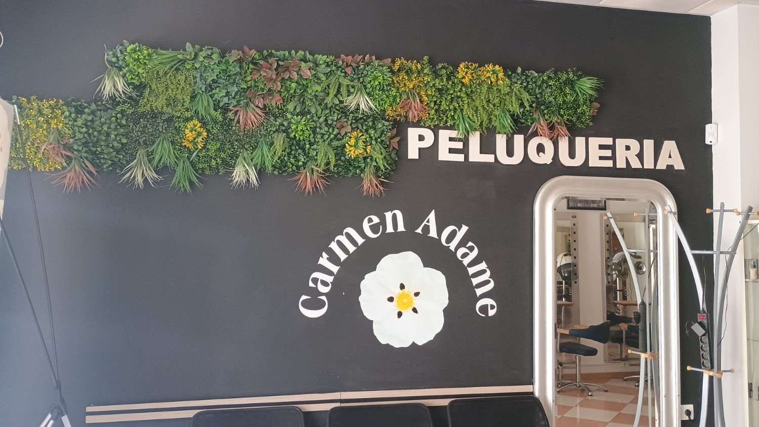 Letrero de salón de belleza con las palabras “PELUQUERIA” y “Carmen Adame” en una pared oscura con decoración vegetal.