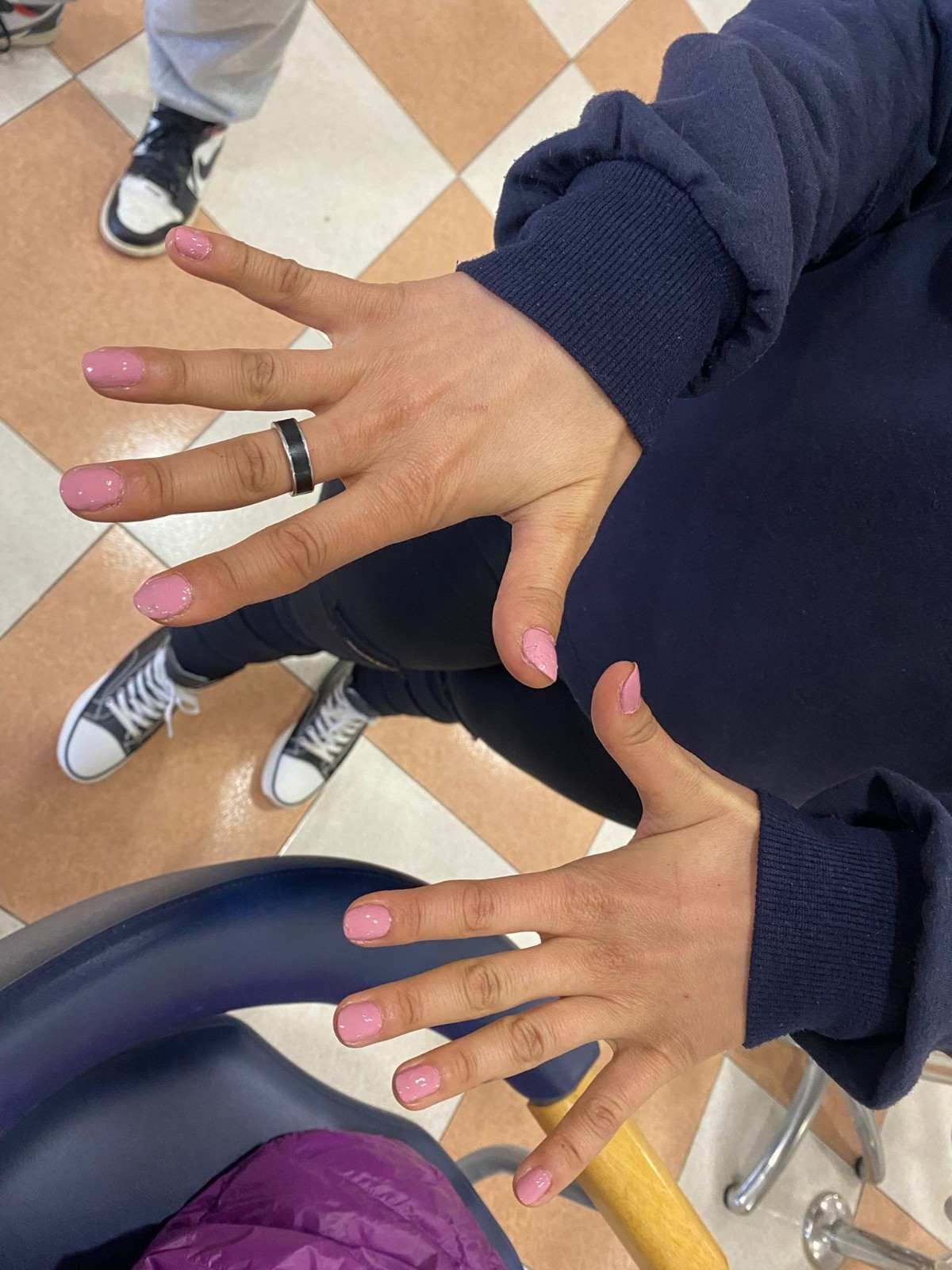 Manos con uñas pintadas de rosa y anillos alzadas sobre un suelo a cuadros en un entorno similar a un aula.