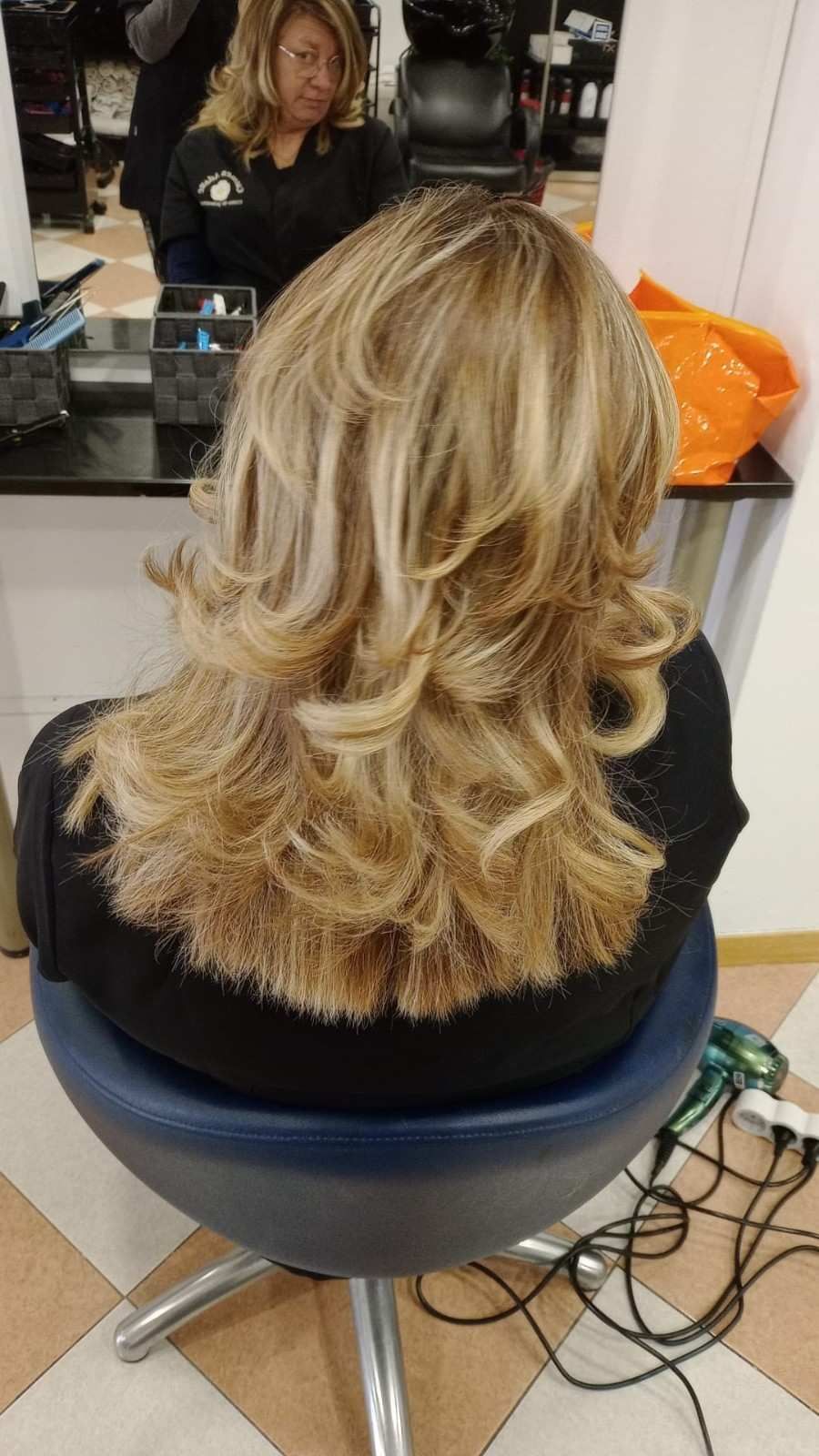 Corte de pelo rubio a capas con puntas rizadas, visto desde atrás en una silla de peluquería con reflejo en el espejo.