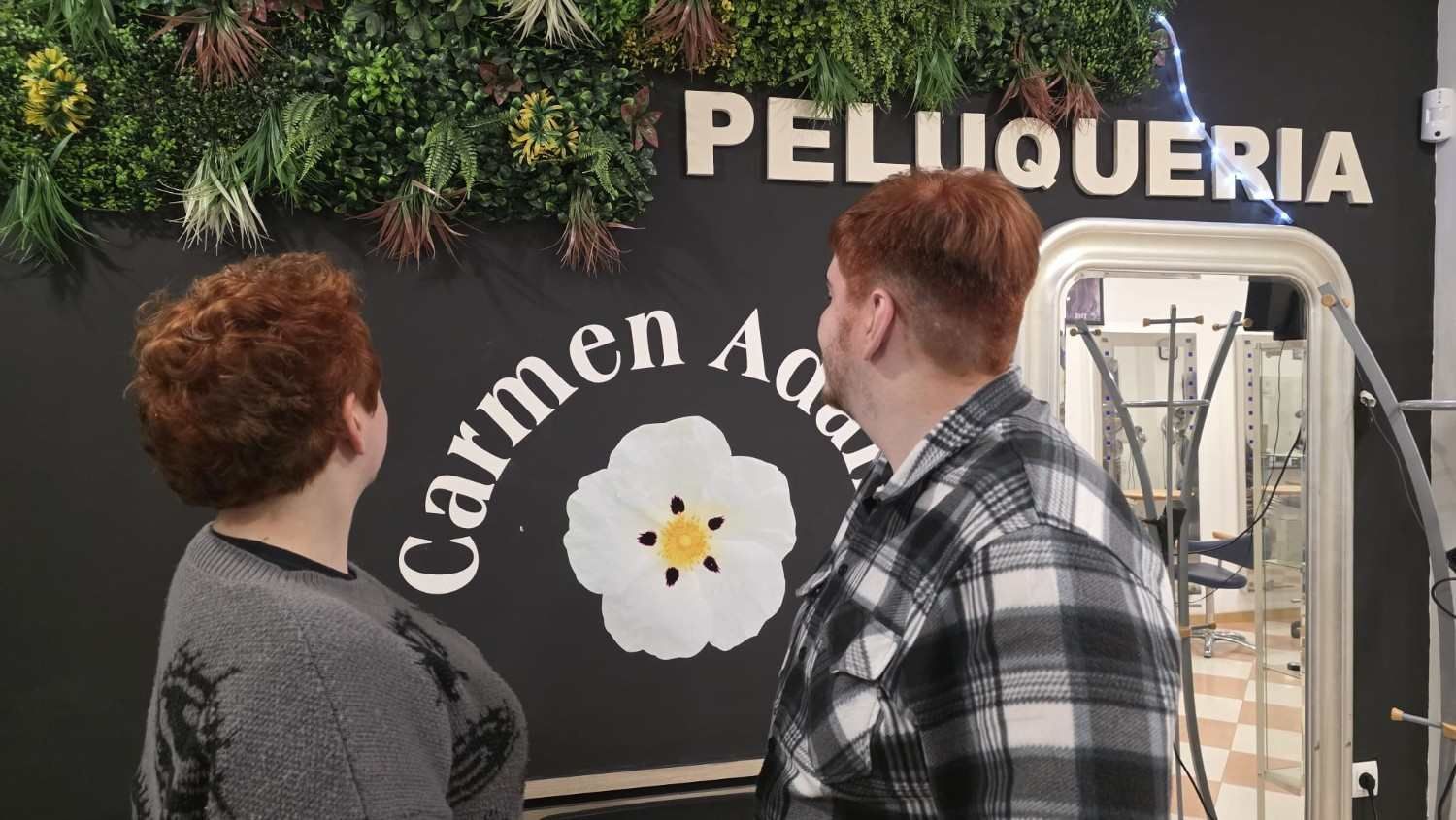 Dos personas mirando un letrero de una peluquería que dice “Peluquería Carmen” con un logotipo de una flor.
