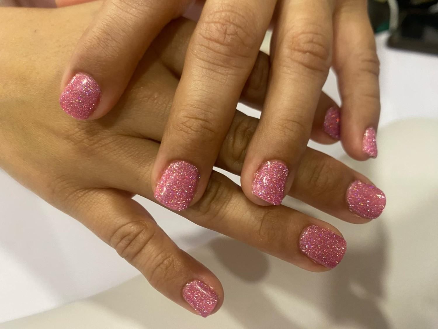 Manos con esmalte de uñas rosa brillante descansando juntas sobre una superficie blanca.
