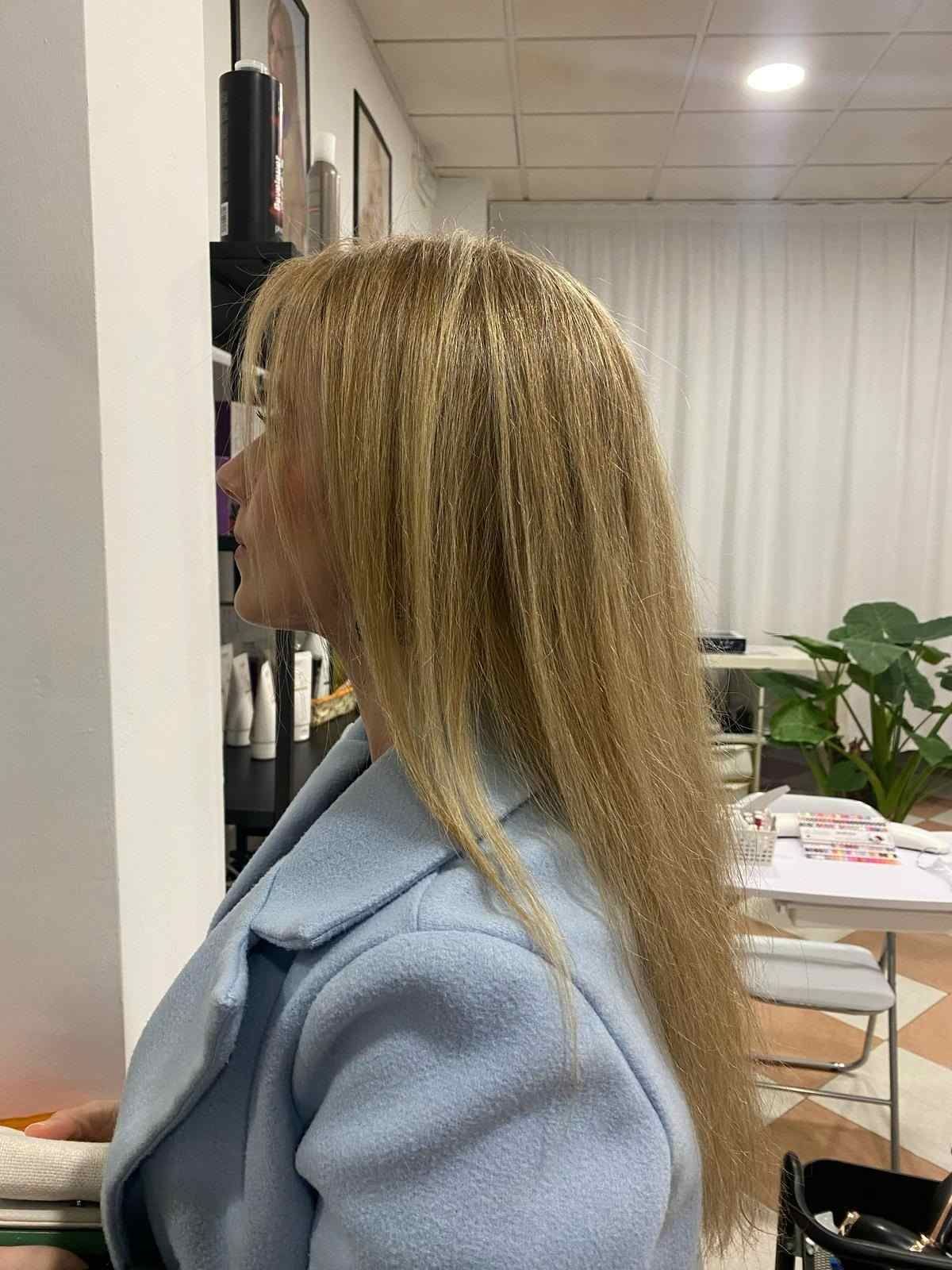 Mujer con abrigo azul claro de pie en el interior de una habitación con plantas y una mesa, junto a una pared.