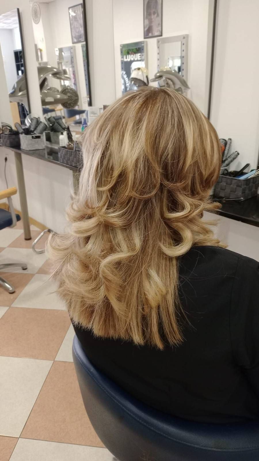 Cabello rubio, con capas y peinado en rizos sueltos, sentado en una silla de peluquería, visto desde atrás.
