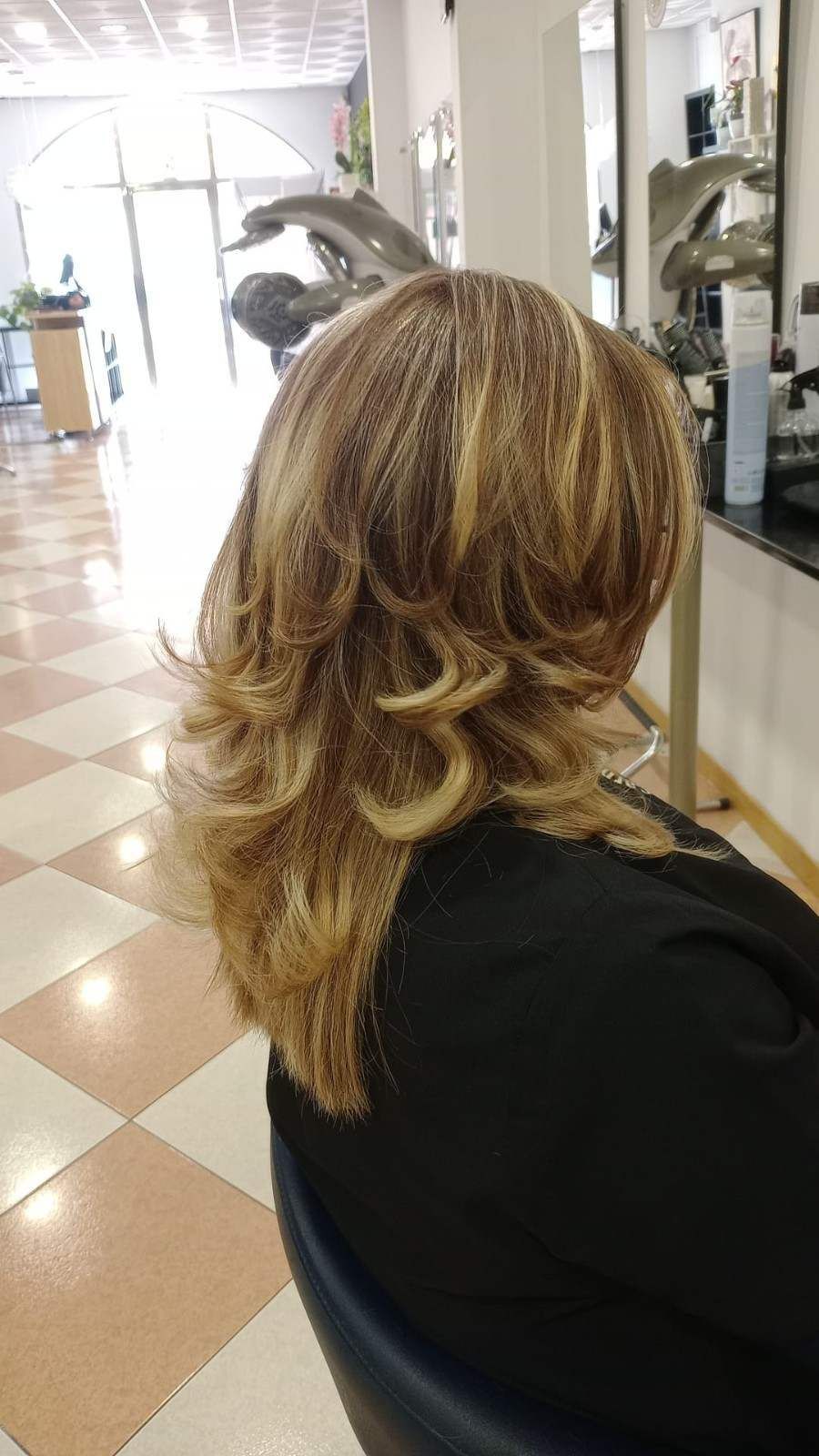 Persona con cabello rubio en capas sentada en una silla de peluquería, vista desde atrás.