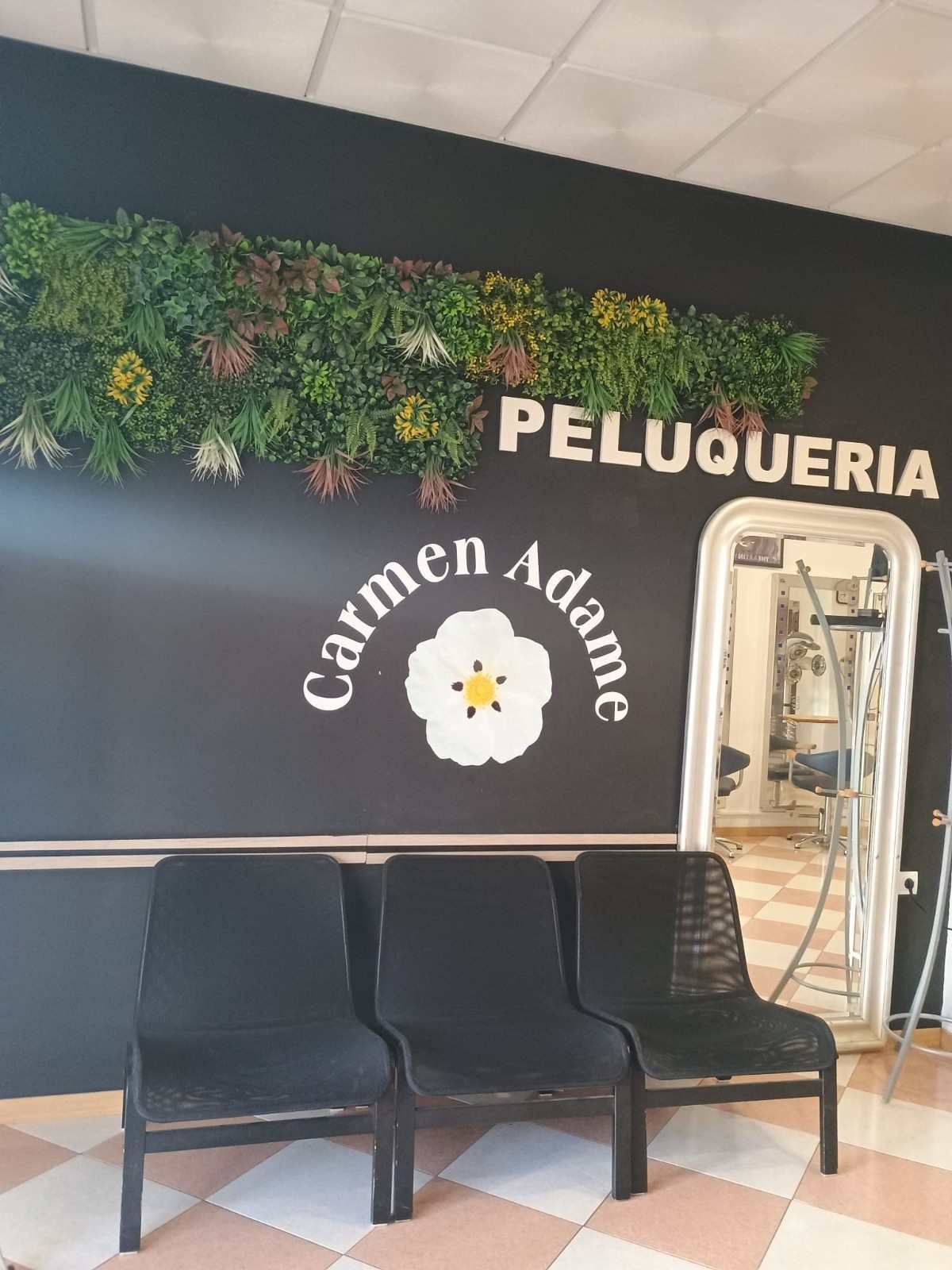 Zona de espera de peluquería con sillas negras, decoración floral en la pared y una puerta de cristal bajo el letrero