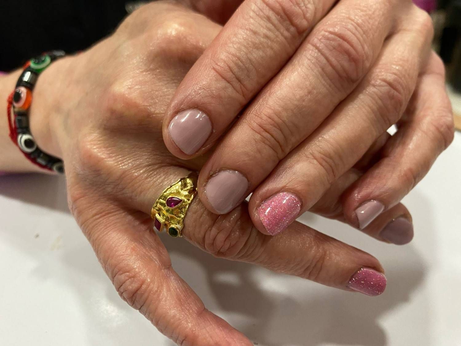 Manos con esmalte de uñas en tonos pastel y rosa con purpurina, luciendo un anillo dorado sobre una superficie blanca.