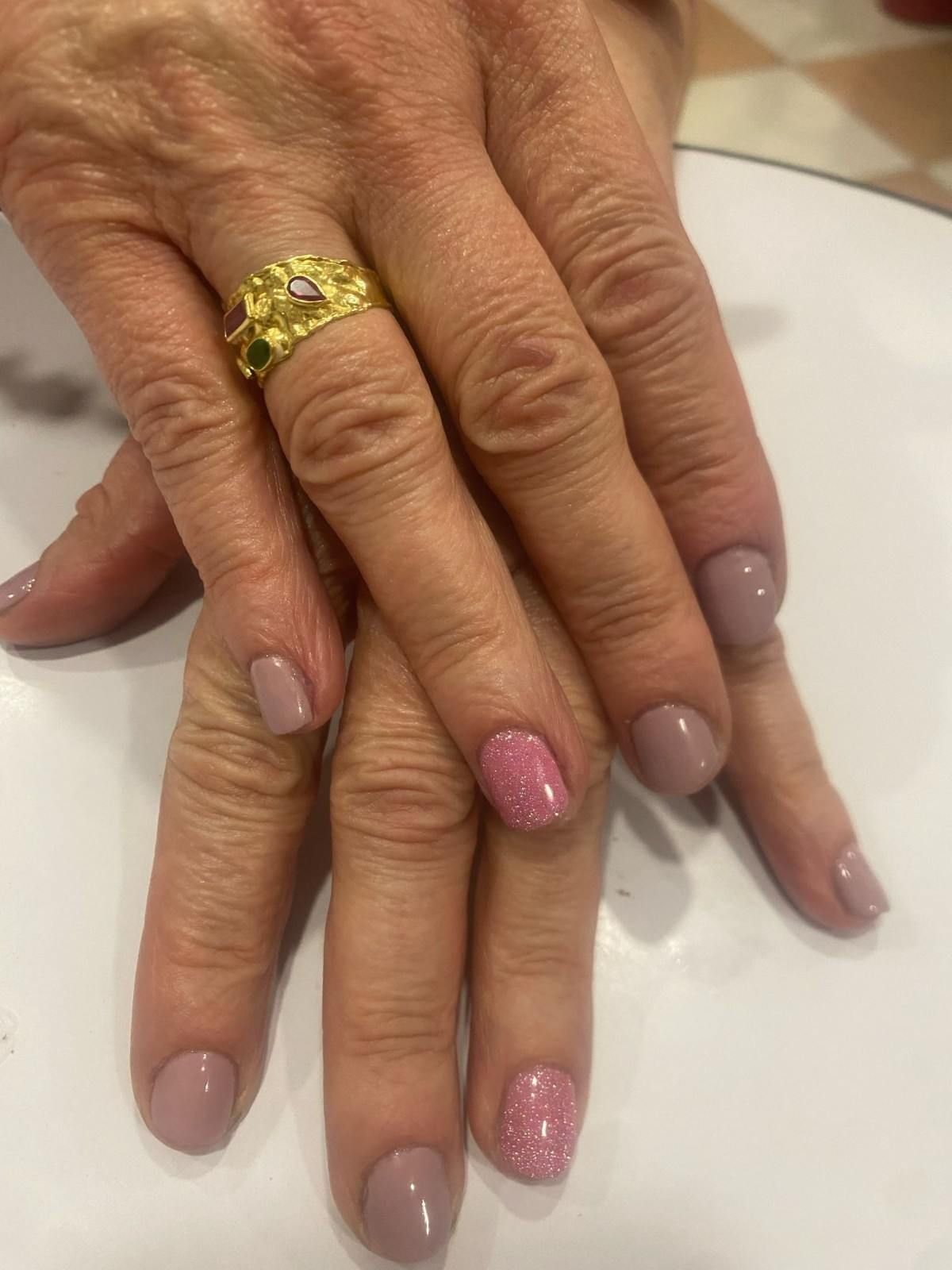Primer plano de manos cruzadas con uñas pintadas en tonos nude y rosa con purpurina y un anillo de oro.