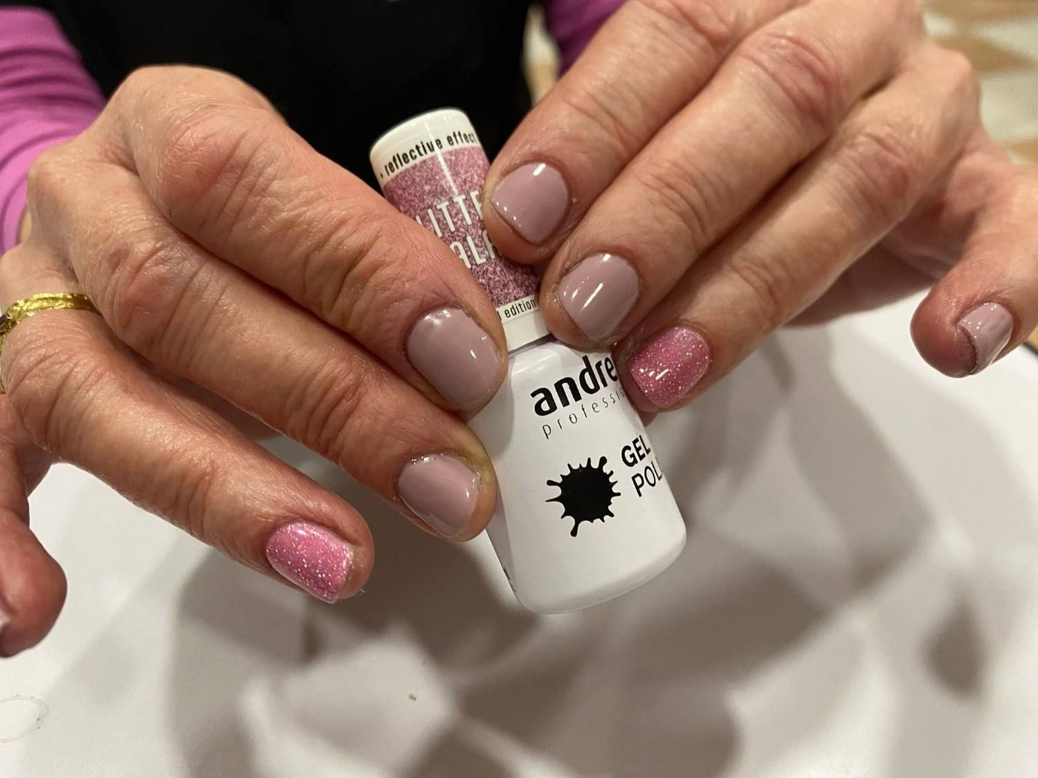 Manos sosteniendo un pequeño frasco de esmalte de uñas con uñas manicuradas en tonos nude y rosa con purpurina.