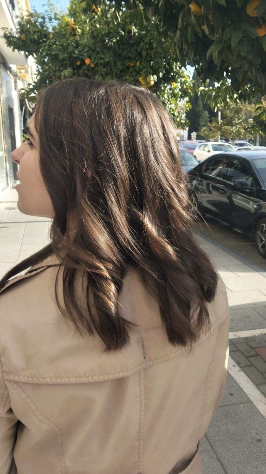 Una persona con cabello castaño ondulado y un abrigo beige está de pie en una acera soleada junto a coches aparcados y árboles.
