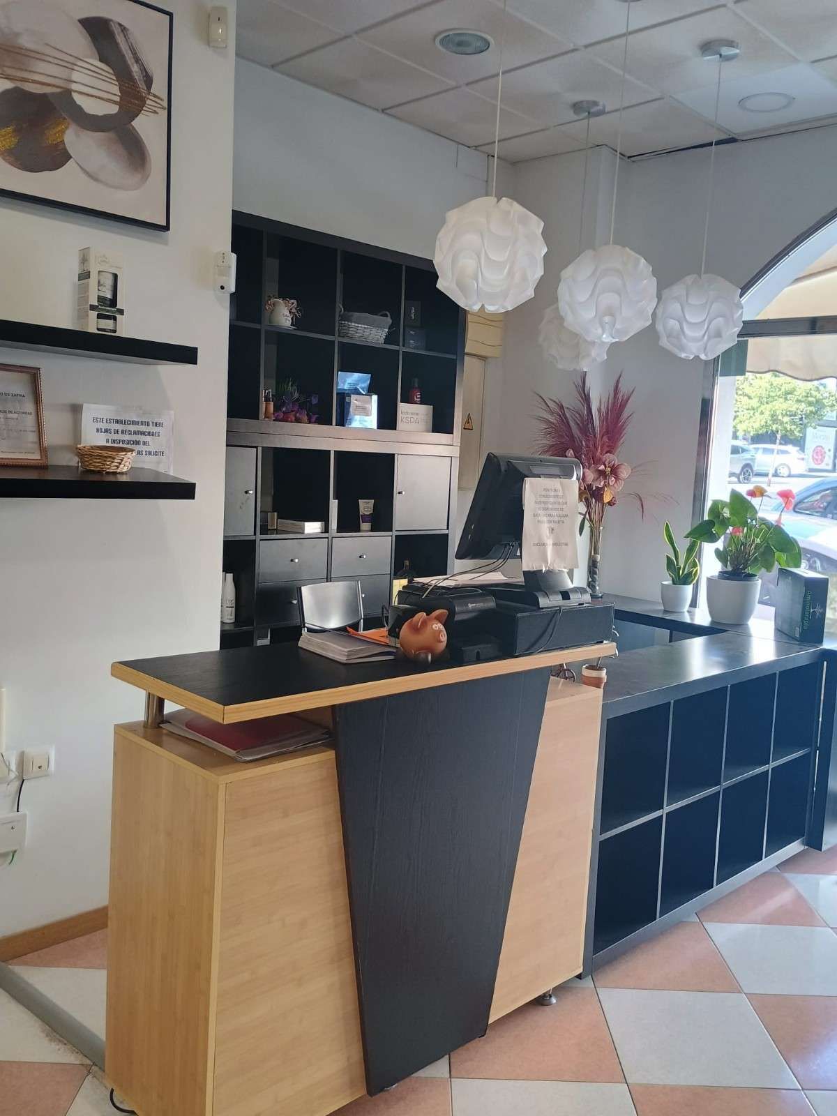 Barra de cafetería moderna con decoración en negro y madera, lámparas colgantes blancas, estantes y una planta en maceta cerca de la ventana.