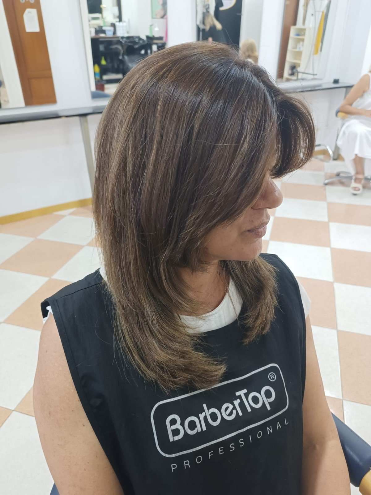 Persona con cabello castaño hasta los hombros, con una capa de peluquería negra, sentada en una peluquería, vista de perfil.