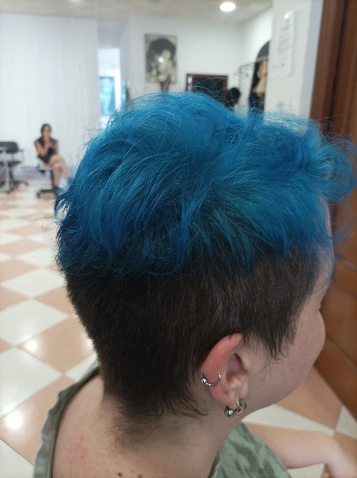 Persona con cabello azul brillante y rapado lateral, vista de espaldas en el pasillo de una peluquería.