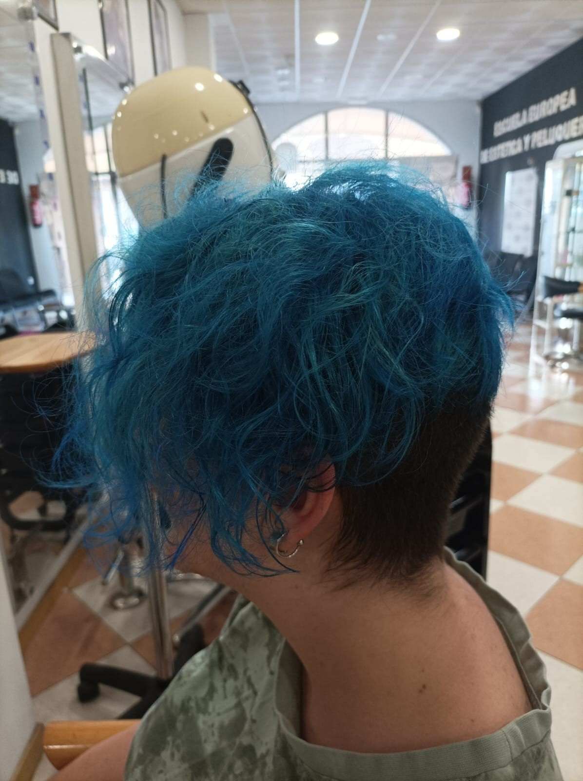 Persona con cabello azul brillante vista de espaldas en el espejo de una peluquería, vistiendo una blusa verde.