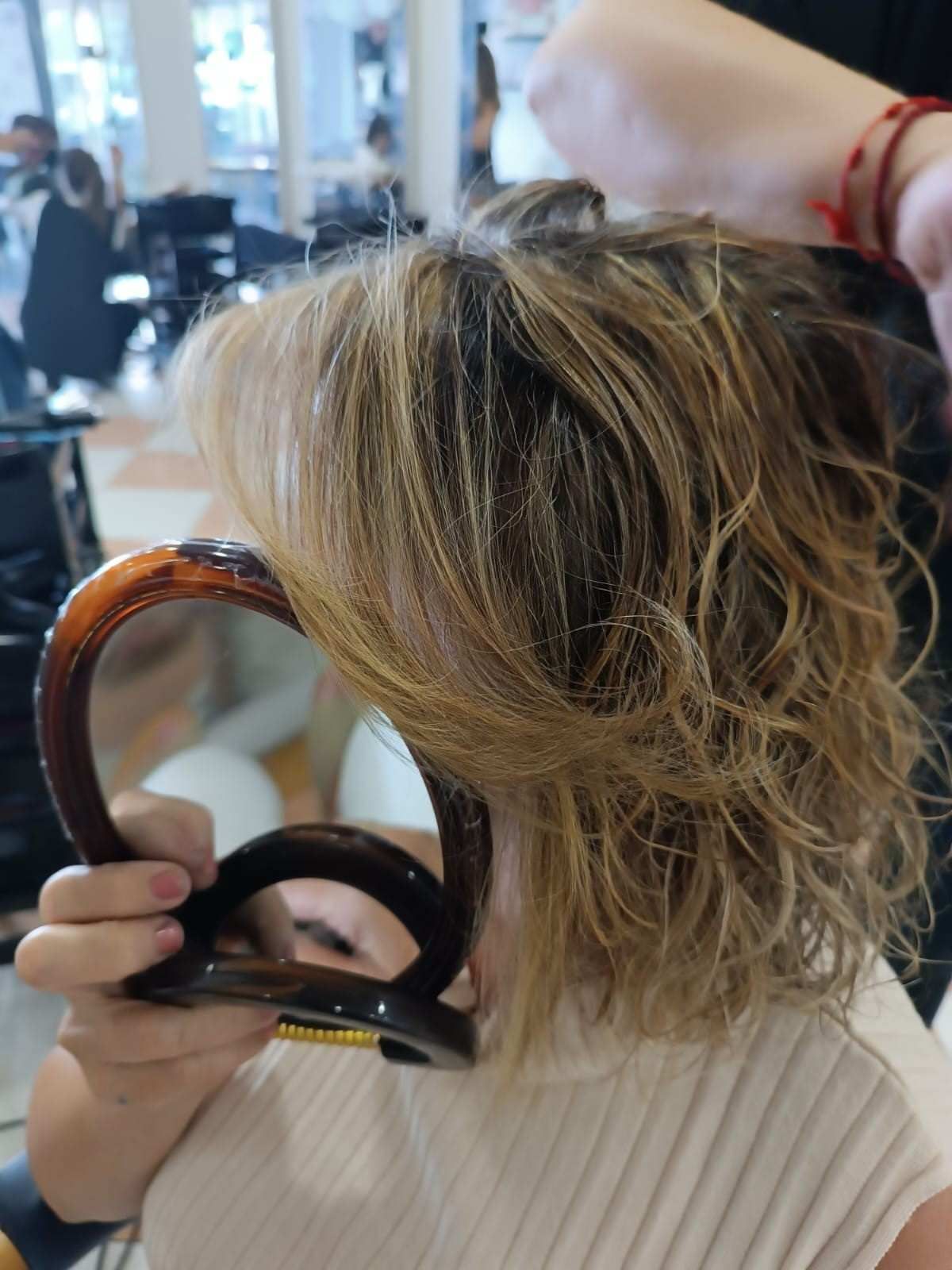 Una persona sostiene un espejo de mano redondo mientras se peina el cabello rubio corto en una peluquería.