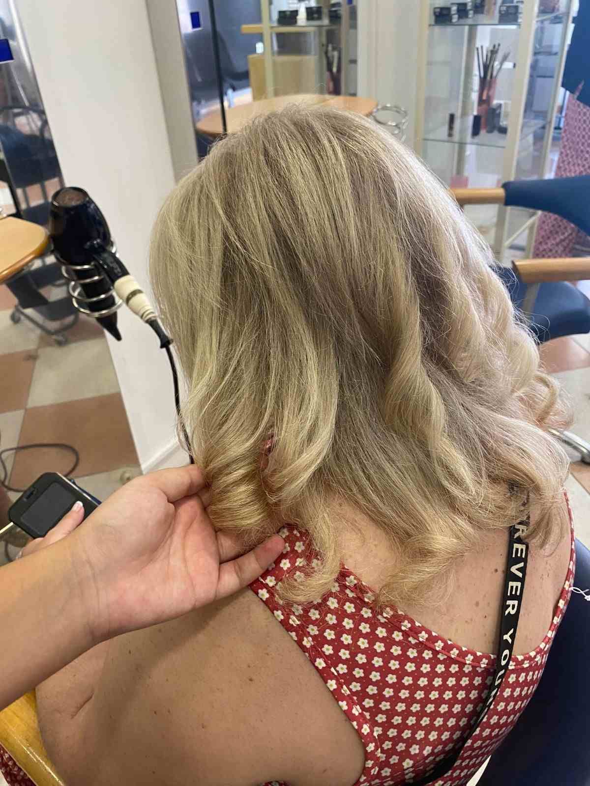 Vista trasera de una persona rubia con una blusa roja de lunares, sentada en una silla de peluquería mientras le peinan el cabello.