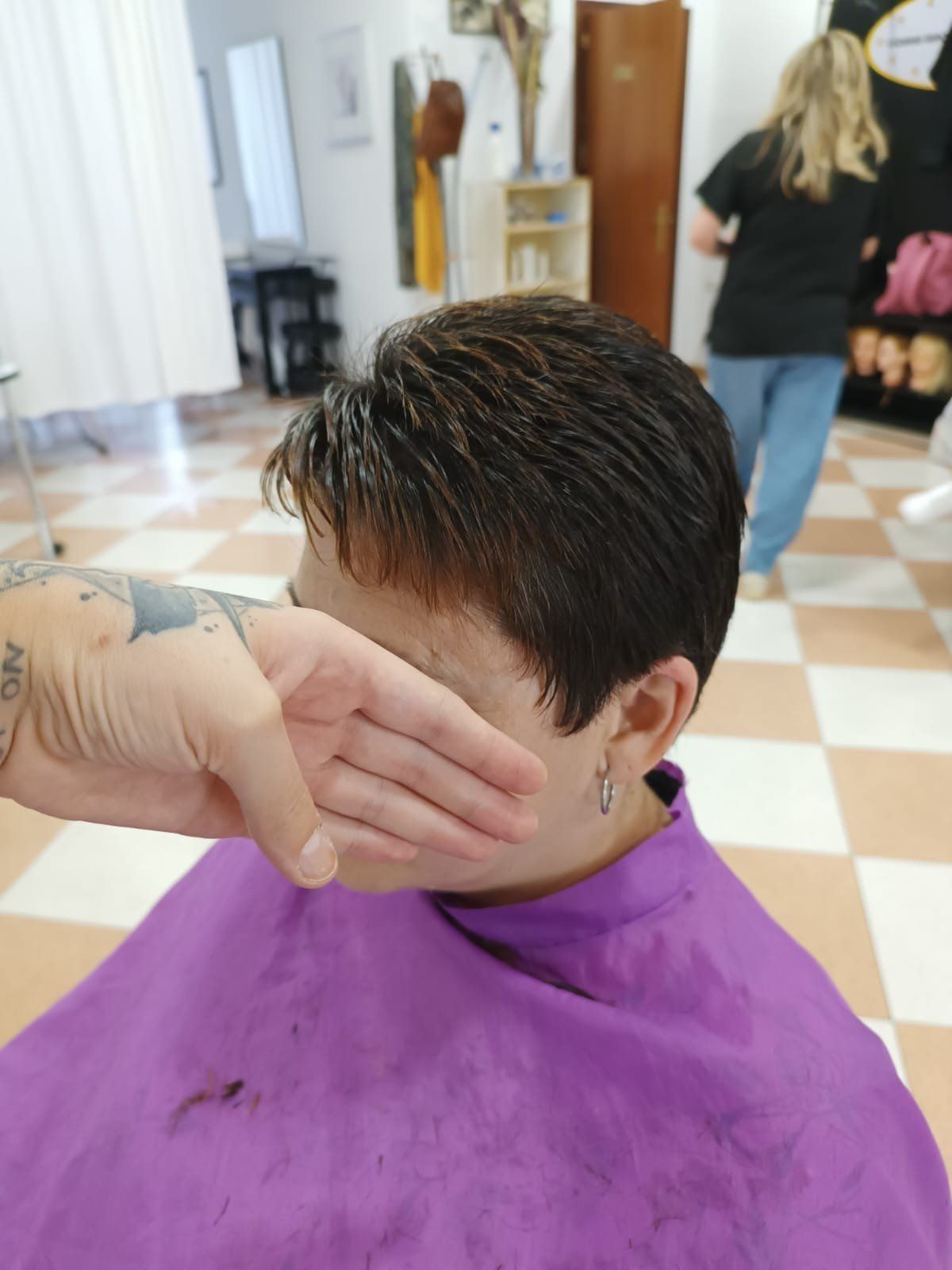 Persona con cabello corto y oscuro cubierto por una capa morada en una peluquería, con la mano cerca de la cara mientras la están arreglando.
