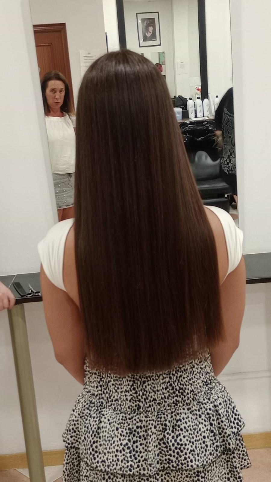 Mujer con cabello largo, liso y castaño, de pie en una peluquería, vista de espaldas, con su reflejo en un espejo.