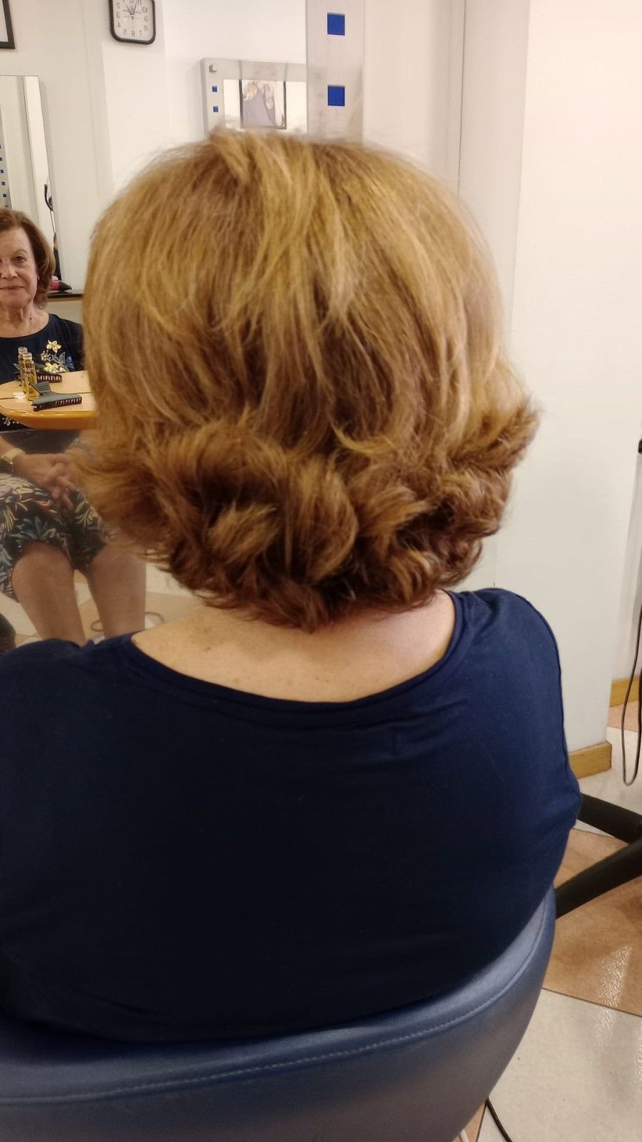 Persona con cabello rubio corto y ondulado sentada en una silla de peluquería, vista de espaldas, con una blusa oscura.