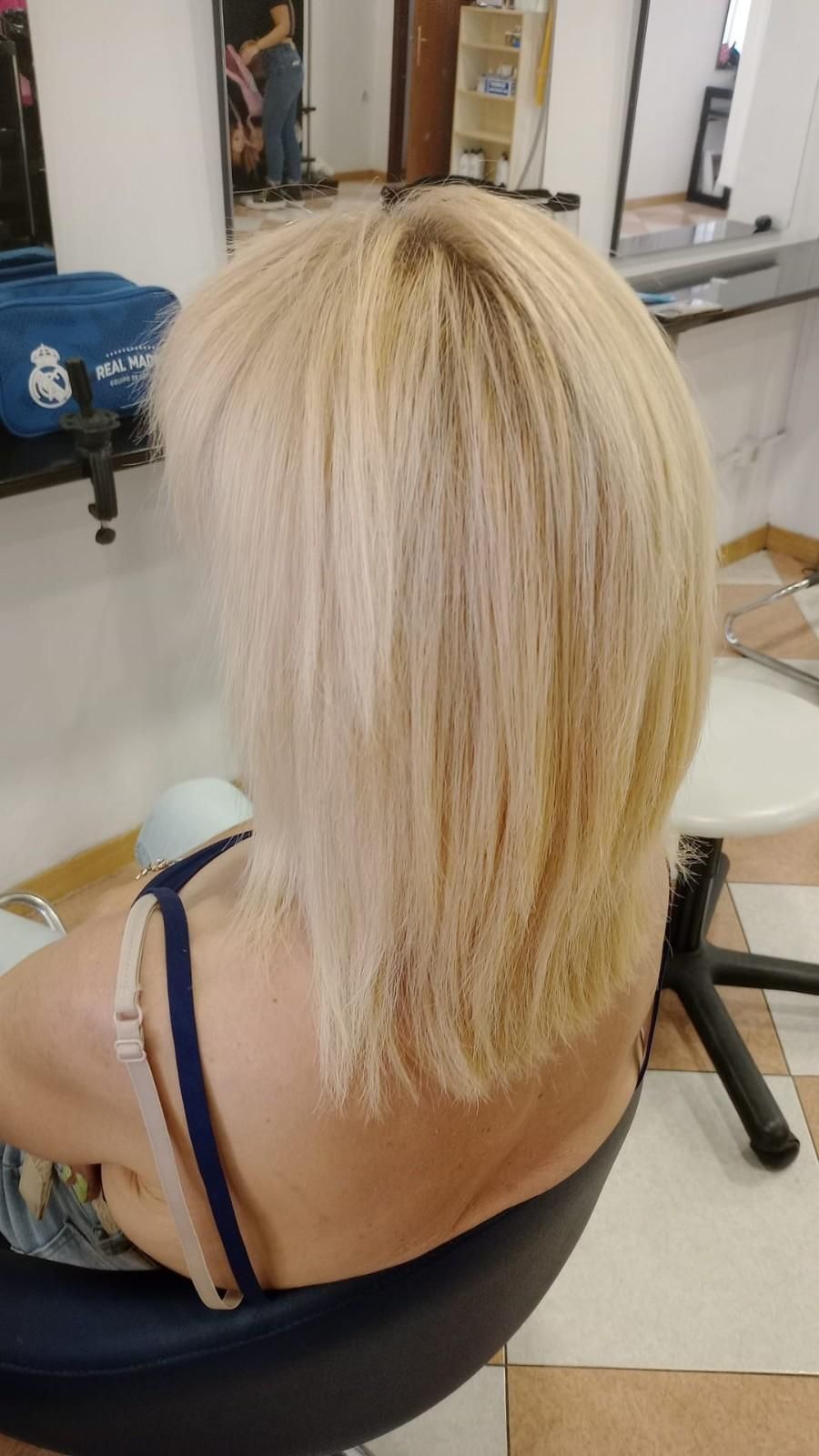 Corte de pelo rubio a capas visto desde atrás en una silla de peluquería, con una tira negra visible sobre el hombro.
