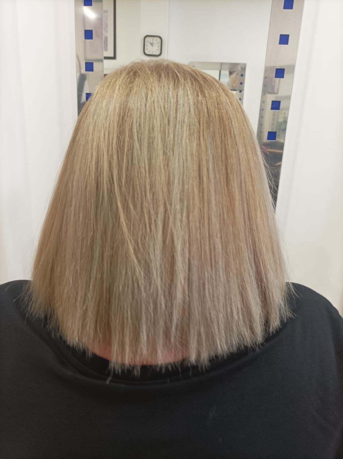 Vista trasera de un corte de pelo bob rubio a la altura de los hombros con un acabado recto y liso, reflejado en el espejo de una peluquería.