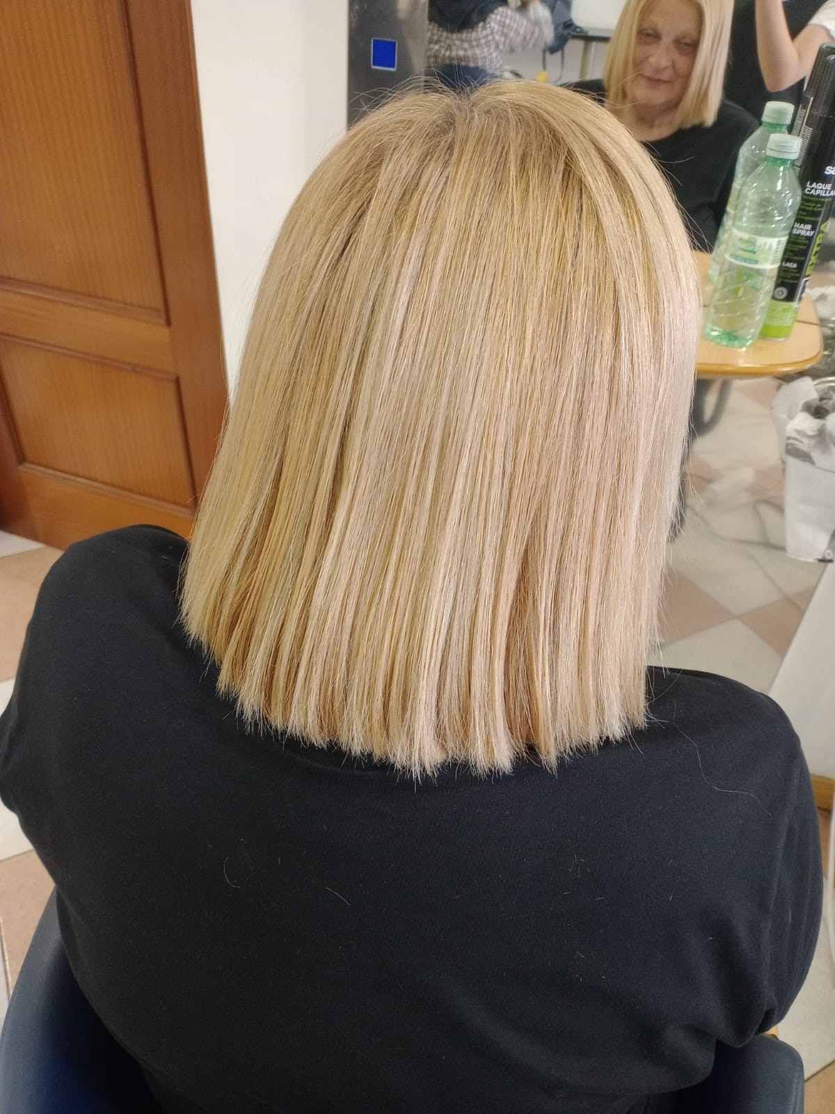 Persona con corte de pelo bob rubio liso, vista de espaldas, con una blusa negra, sentada en el interior cerca de una mesa con botellas.