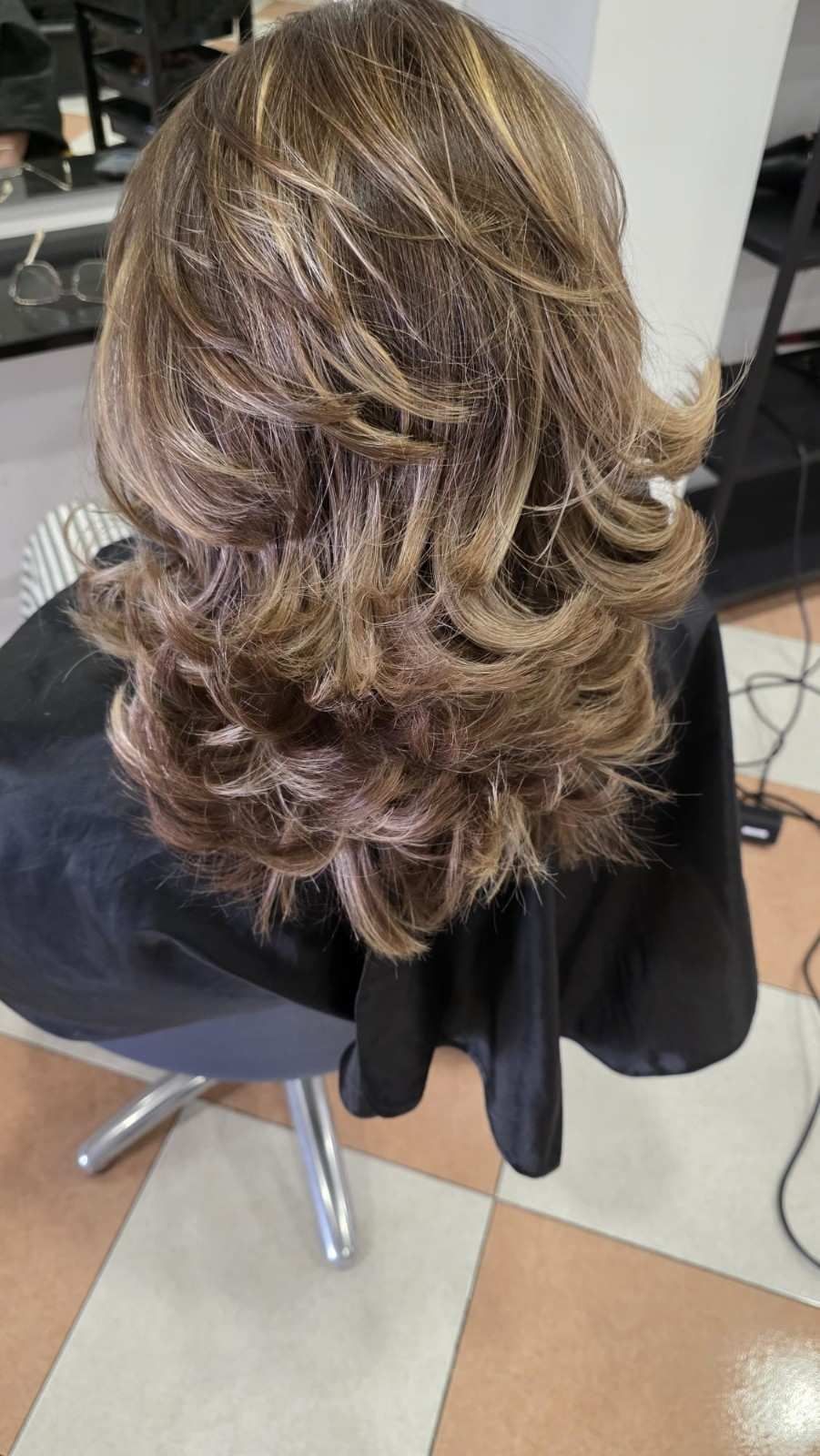 Cabello ondulado, castaño y rubio, con capas, visto desde atrás, con una capa negra de peluquería en una silla de salón.