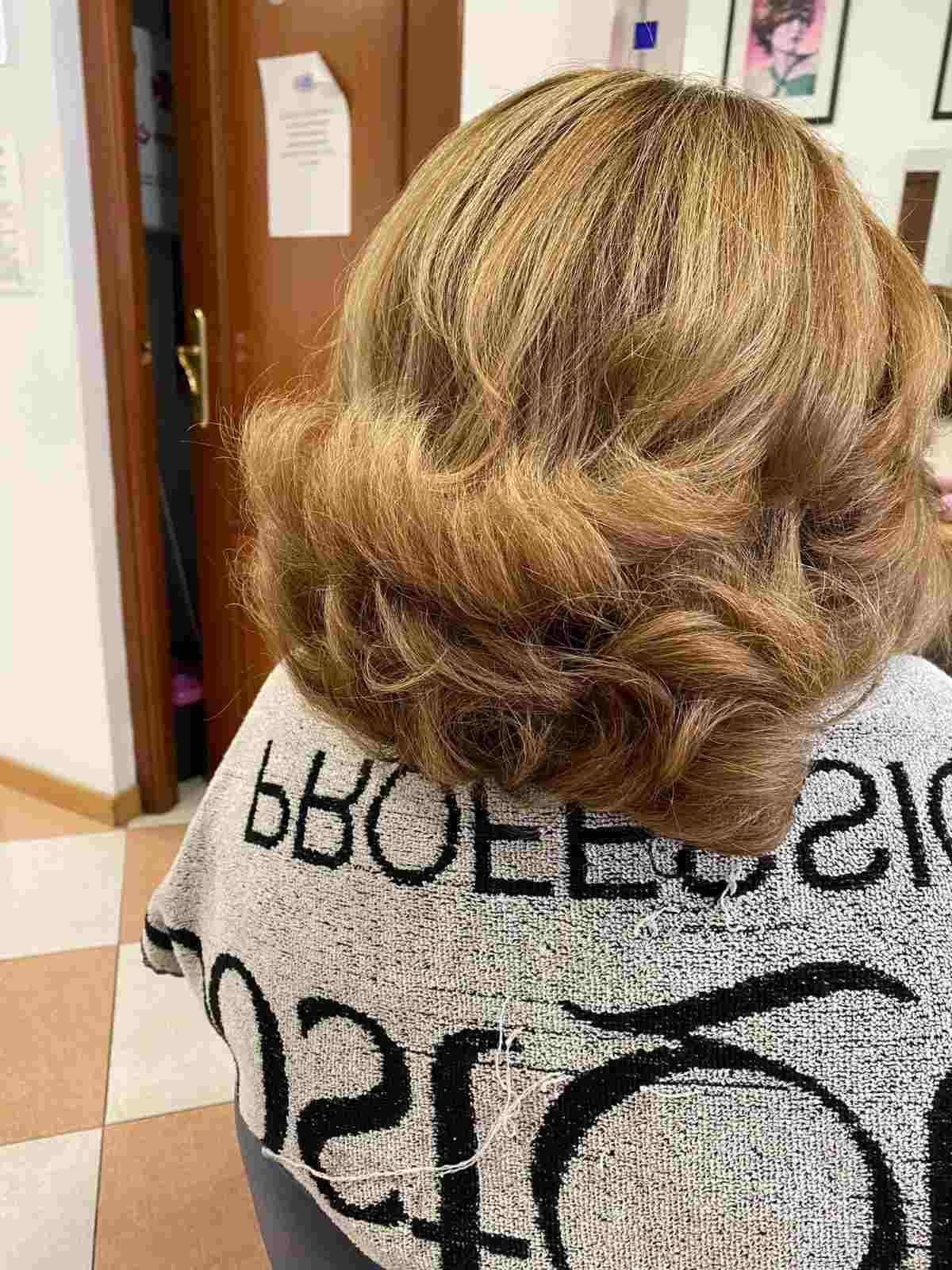 Persona con cabello rubio hasta los hombros vista de espaldas en una habitación, vistiendo una camisa gris con texto negro.