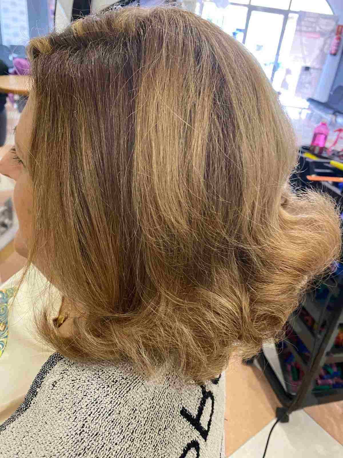 Vista lateral de una melena rubia-castaña capeada hasta los hombros con suaves rizos en una silla de peluquería.