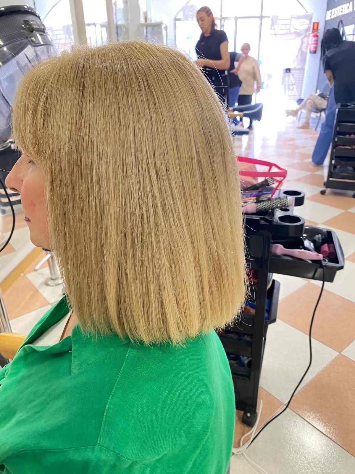 Corte de pelo bob rubio con puntas rectas en una peluquería; la clienta lleva una capa verde.