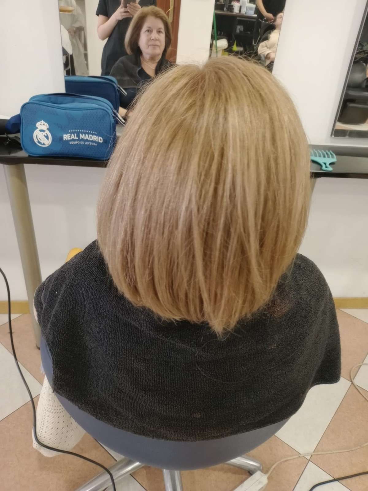 Vista trasera de una persona rubia sentada en una silla de peluquería, frente a un espejo, con una bolsa azul cerca.