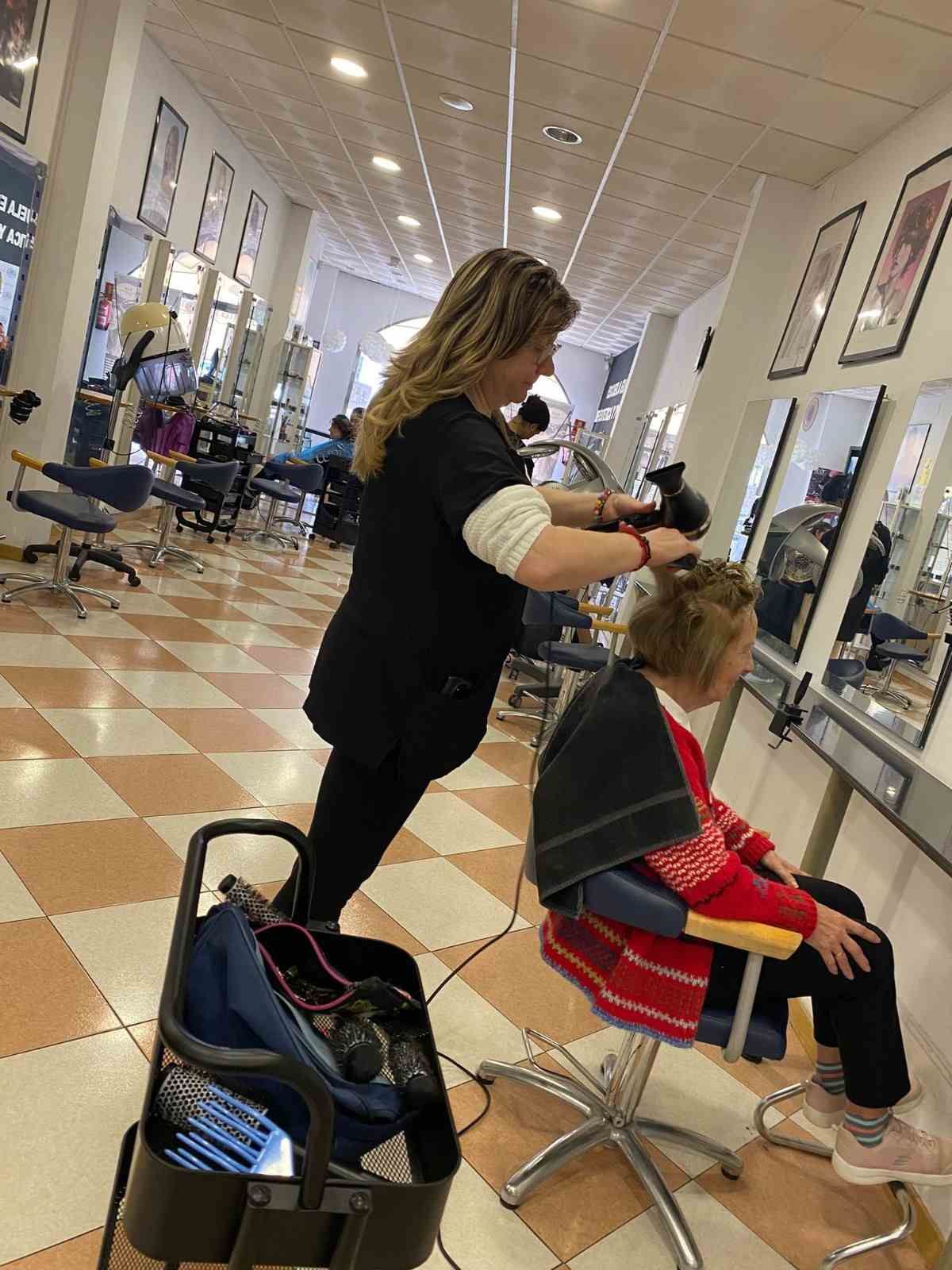Una estilista le corta el pelo a una clienta en un salón luminoso, con espejos, sillas y suelo de baldosas.