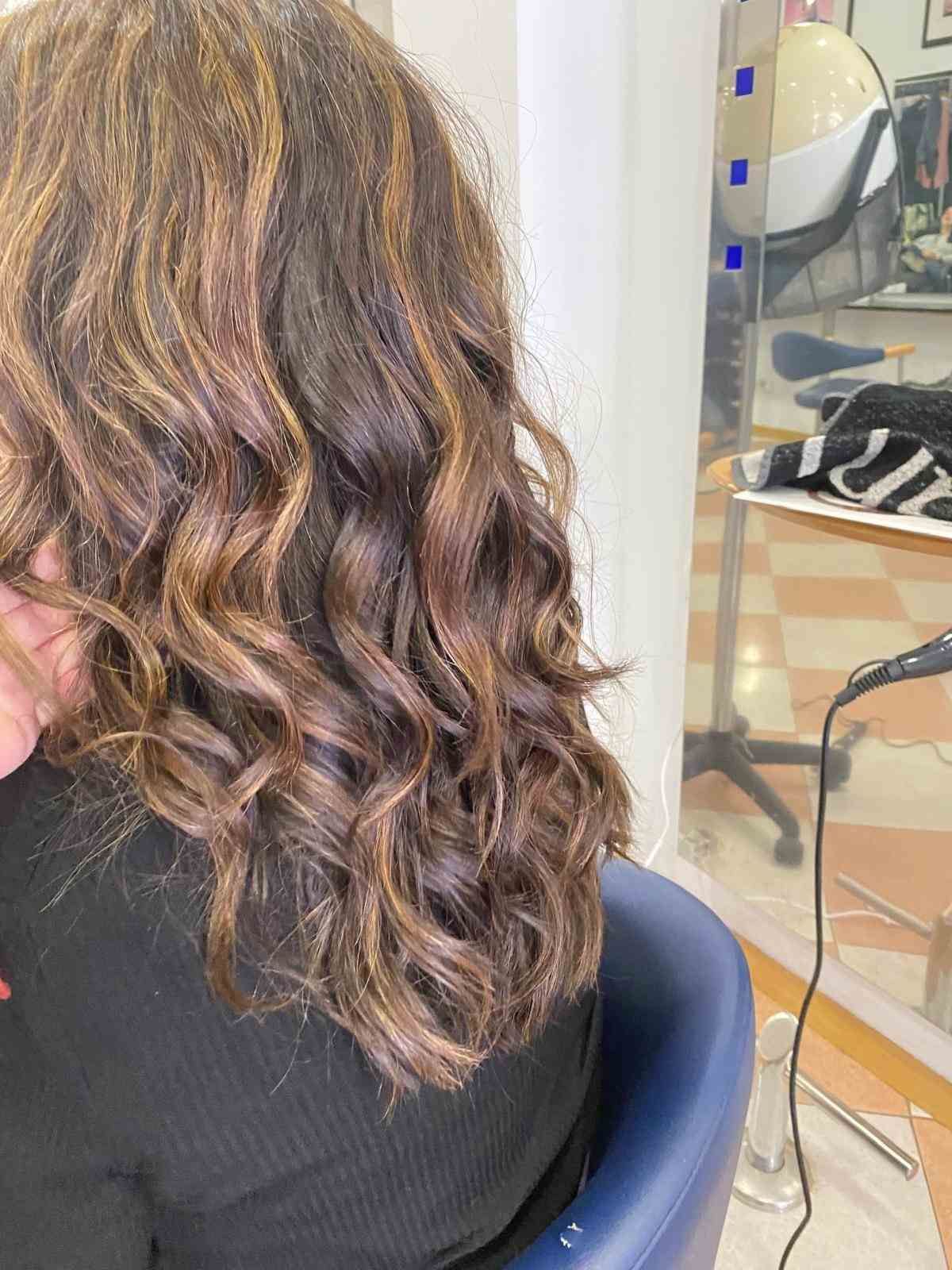 Cabello castaño rizado con reflejos rubios, visto desde atrás en una silla de peluquería.