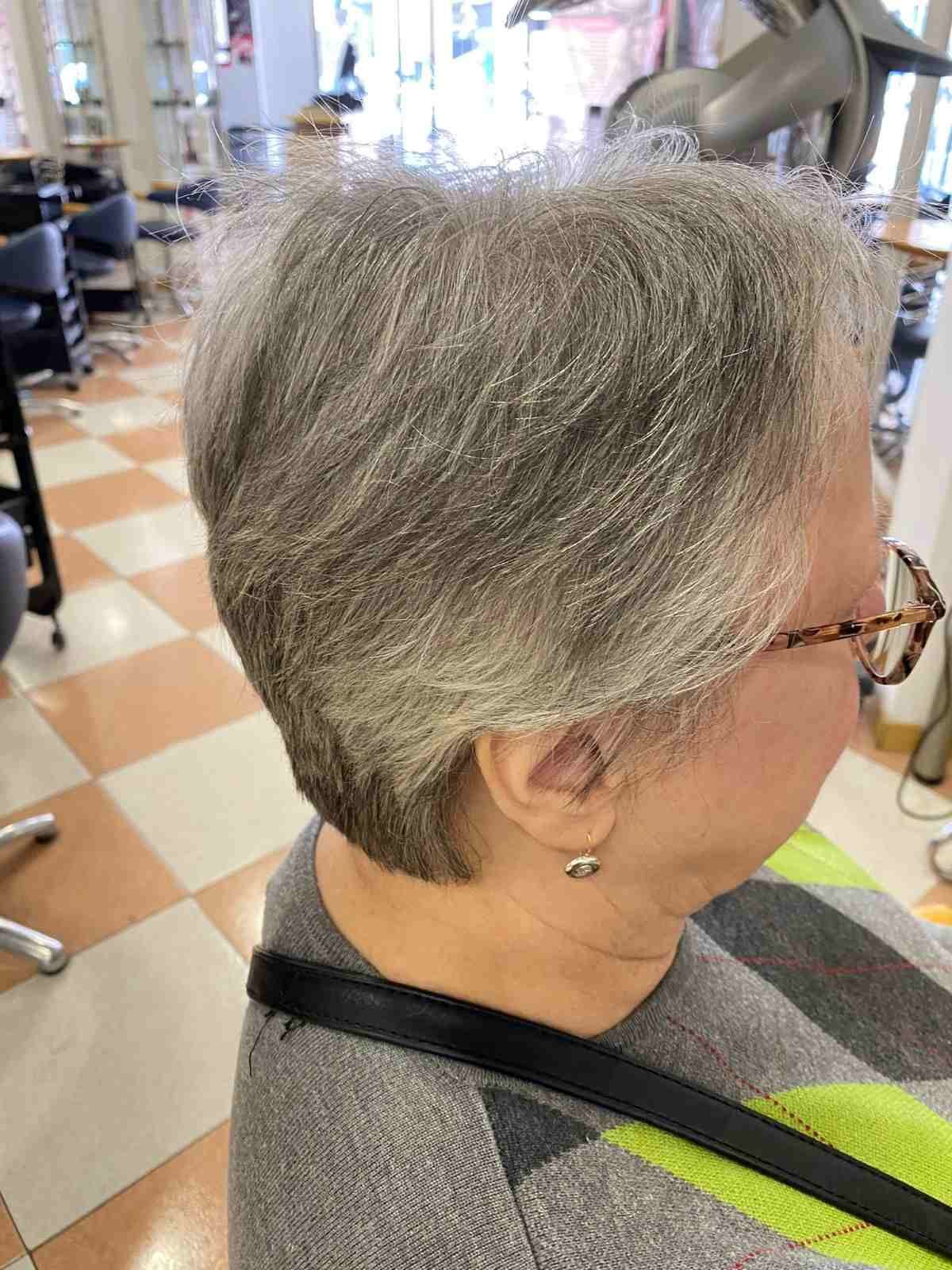 Perfil de una persona con cabello corto y gris y gafas, sentada en una silla de peluquería, en el interior de un edificio con suelo de baldosas a cuadros.