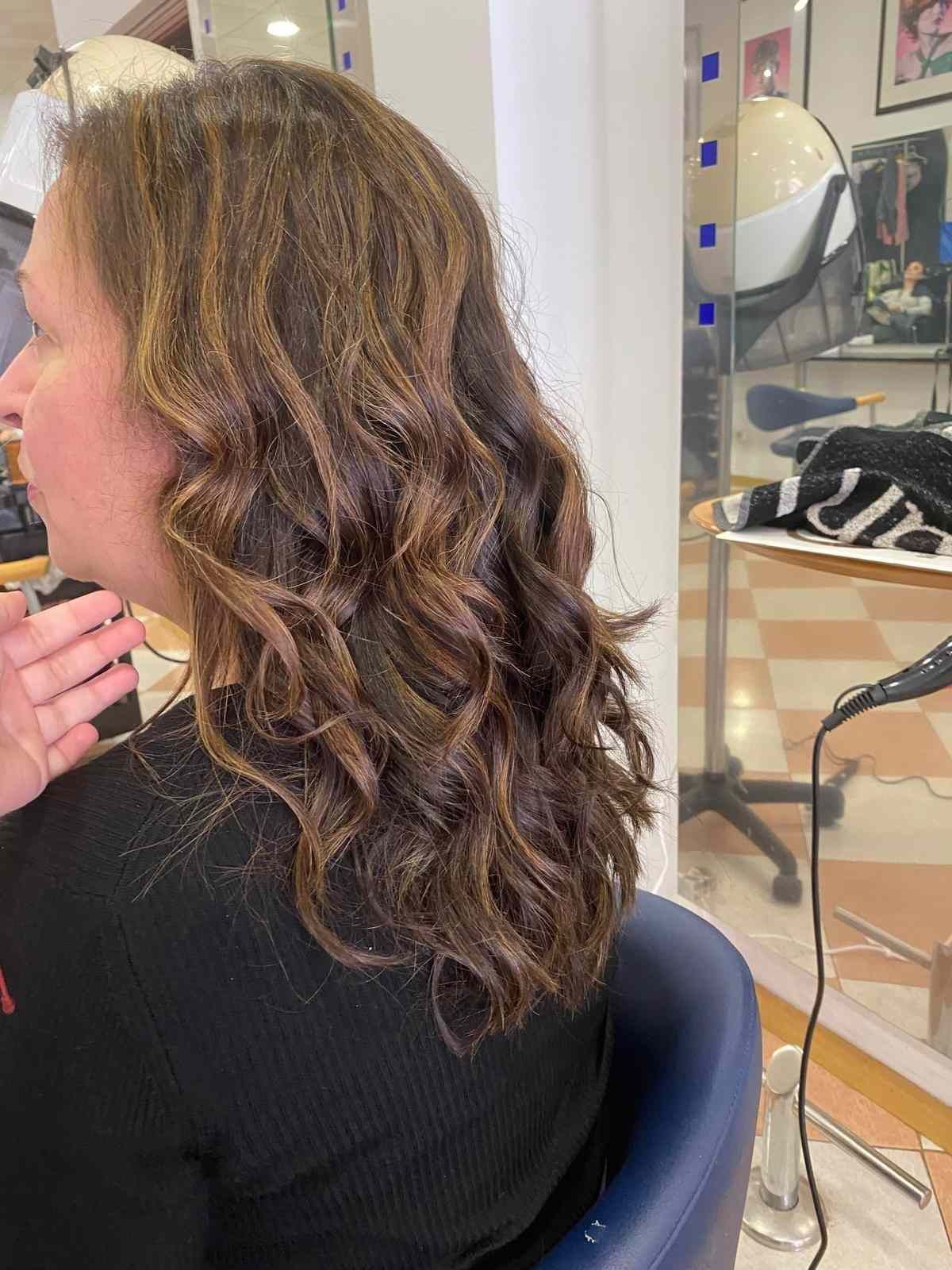 Vista lateral de una persona con cabello castaño rizado sentada en una silla de peluquería mientras le peinan.