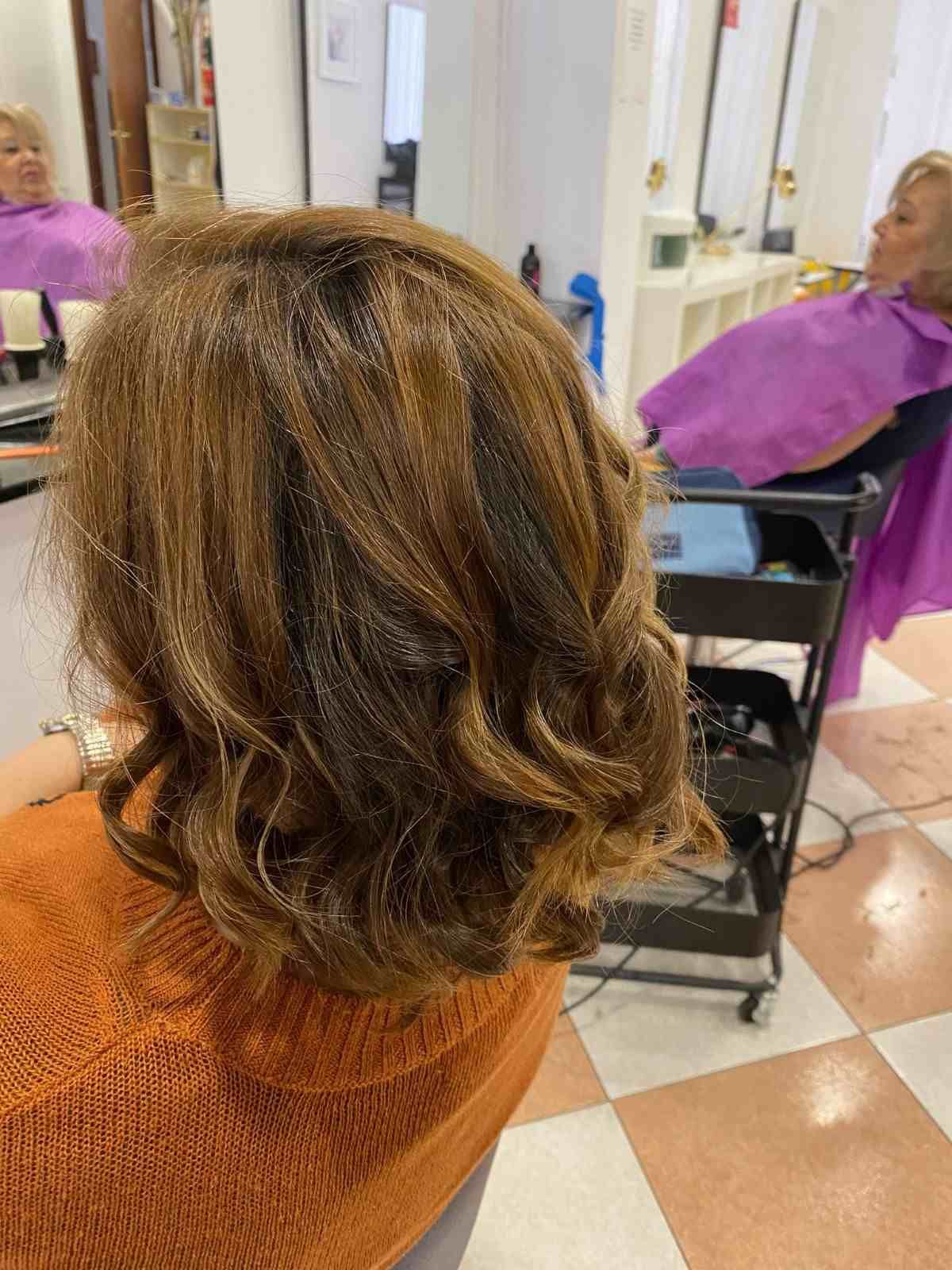 Clienta de peluquería con cabello castaño hasta los hombros, vestida con una blusa naranja, sentada en una silla de salón con espejos.