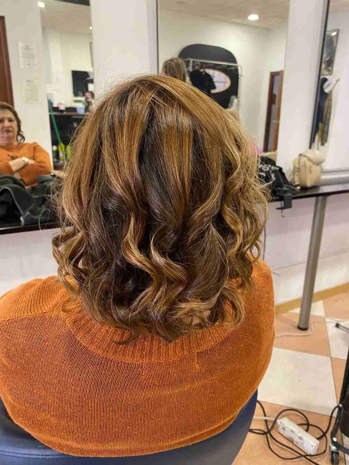 Mujer con cabello castaño ondulado hasta los hombros sentada en una silla de peluquería, vista desde atrás.