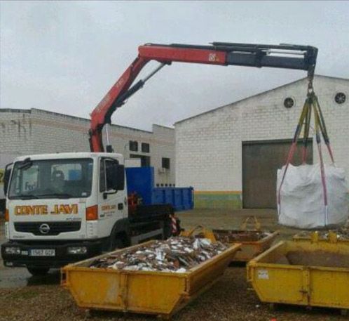 Camión con grúa levantando una gran bolsa, contenedores de chatarra frente al almacén.
