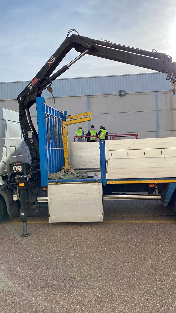 Camión con grúa cargando grandes paneles blancos; tres trabajadores con chalecos supervisan el proceso cerca de un almacén.