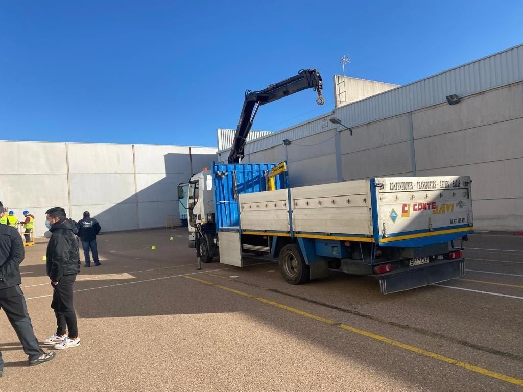 Camión con grúa extendiéndose hacia arriba; gente parada cerca en un área pavimentada, bajo un cielo azul.