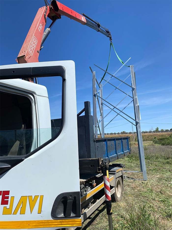 Camión con grúa levantando un marco de metal contra un cielo azul, al aire libre.