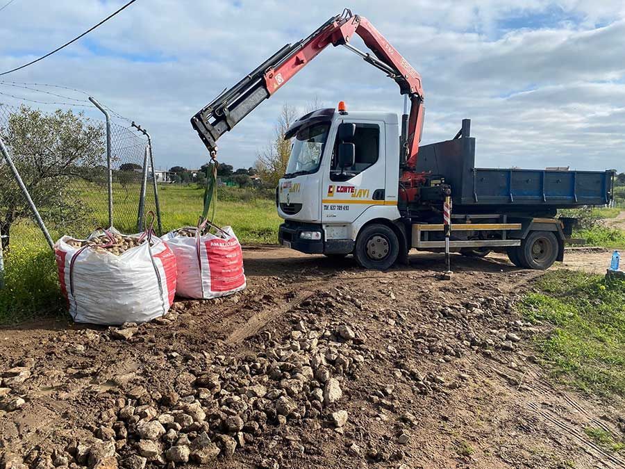 Camión grúa cargando bolsas de escombros en un camino de tierra, cielo nublado.