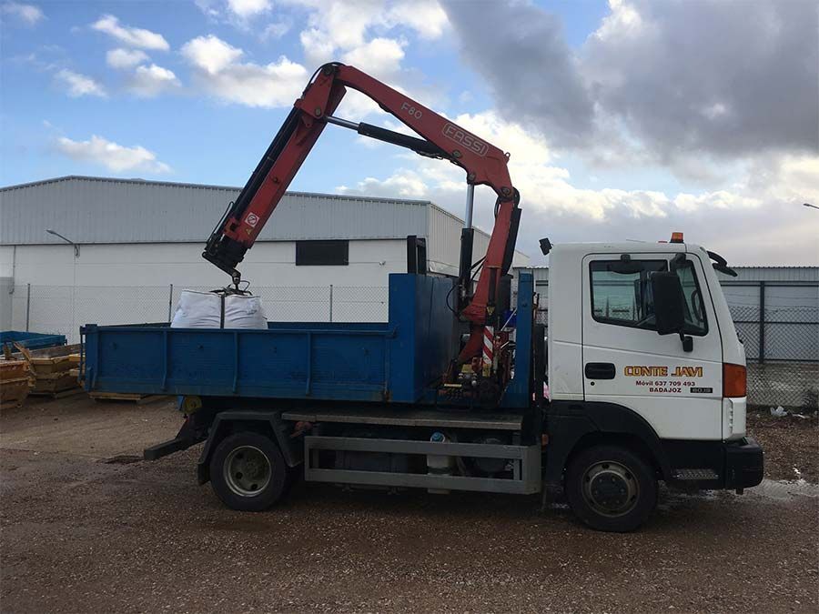 Un camión con una grúa roja levanta una bolsa de una caja azul bajo un cielo nublado.