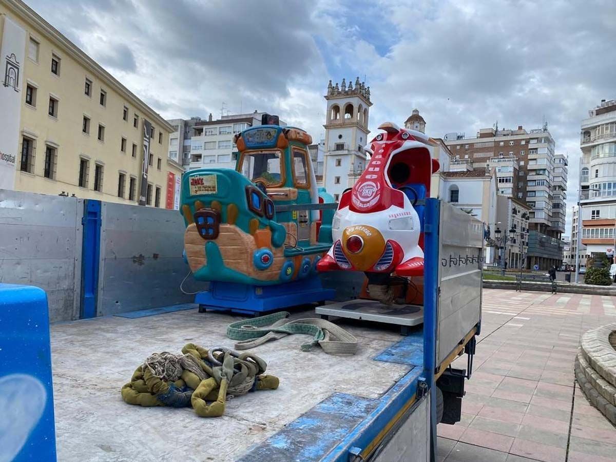 Un tren de juguete y un cohete viajan en la caja de un camión en una plaza de la ciudad, con edificios al fondo.